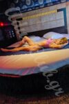 Woman wearing pink lingerie reclining on a round white bed in a dark, modern bedroom.