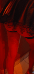 Dim red lighting shows a person standing, capturing their lower torso and legs in a short, lacy skirt with visible tattoos on the thighs near a tiled floor.
