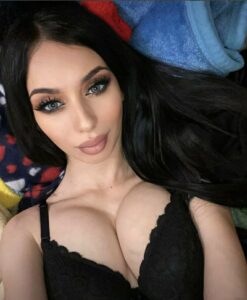Selfie of a woman with long dark hair wearing a black lace bra, looking at the camera.