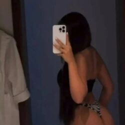Person taking a mirror selfie in a dark room, wearing a black bikini top and zebra-pattern bikini bottoms, with long dark hair and holding a white smartphone to cover their face.