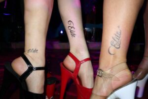 Close-up of three ankles with 'Elite' tattoos, each wearing a different high heel (black, red, and clear-beige) in a nightclub setting.