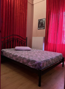 Metal-framed bed with a floral mattress and a single pillow in a red-curtained bedroom.