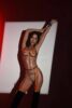 Woman in orange strappy lingerie posing with arms raised against a white panel in a dark studio, red-tinted lighting. Woman in orange strappy lingerie posing with arms raised against a white panel in a dark studio, red-tinted lighting.