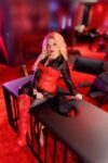 Woman in a red corset and red knee-high boots sits on a black bar counter in a dim red-lit nightclub, leaning on her hand.