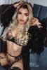 Busty woman with long blonde curls in black lace lingerie and a feathered robe posing on a sofa, seductive pose.