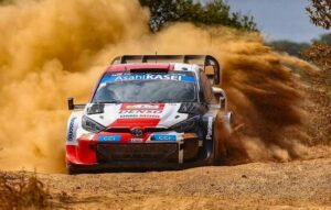 Rally car speeding on a dirt road, spraying a large orange dust cloud behind it; white, red, and blue livery with sponsor logos visible on the car's body.