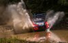 Red rally car plows through a shallow water crossing, spraying muddy water in both directions with a reflection visible in the wet ground.