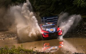 Red rally car plows through a shallow water crossing, spraying muddy water in both directions with a reflection visible in the wet ground.