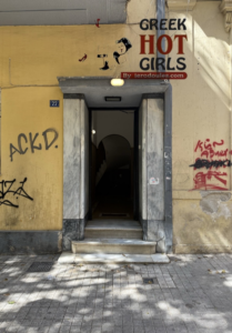Open doorway framed by stone columns on a graffiti-covered yellow building, with a 'GREEK HOT GIRLS' ad above."