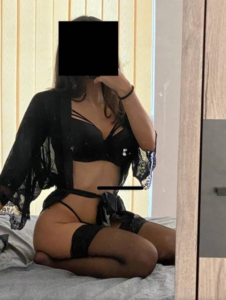 Person in black lingerie posing for a mirror selfie on a bed; face is covered with a black square.