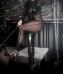 Person in a sheer black catsuit and thigh-high boots sits on a padded bench in a dungeon-like room, holding a whip.