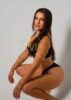 Model posing in a black bikini against a light backdrop, kneeling with long dark hair and visible tattoos; watermark reads 'VIP Greece Escorts'.