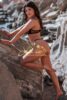 Woman in a black lace bikini poses on rocky shoreline, facing the camera; watermark reads 'Girls Athens Escorts' across the image.