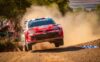 Red rally car jumps over a dirt road, kicking up a large dust cloud with spectators watching from the side.