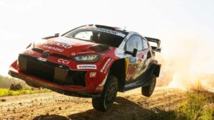 Red rally car is airborne over a dirt road as dust flies, mid-race action on an off-road course.