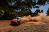 Red rally car speeding along a dirt trail, kicking up a cloud of dust behind it in a forested area.