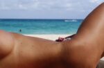Person sunbathing on a sandy beach with turquoise sea and horizon in the background.
