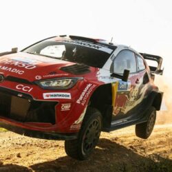 Red rally car airborne over a dirt track, dust spraying behind it.