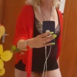 Person taking a mirror selfie in a red cardigan over black lingerie, with neon green nails and white earphones visible at the phone.