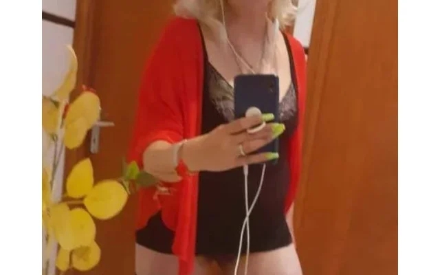 Person taking a mirror selfie in a red cardigan over black lingerie, with neon green nails and white earphones visible at the phone. Person taking a mirror selfie in a red cardigan over black lingerie, with neon green nails and white earphones visible at the phone.