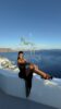 Promotional image of a woman in a black dress and heels posing on a white terrace with the Aegean Sea in the background and a VIP Greece Escorts watermark.
