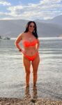 Woman in a bright orange bikini standing in shallow water at a beach, mountains in the distance and a boat on the lake nearby.