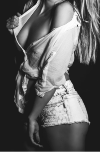 Black-and-white photo of a woman standing sideways in a partially unbuttoned white shirt and distressed denim shorts, against a dark backdrop.