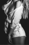 Black-and-white photo of a woman standing sideways in a partially unbuttoned white shirt and distressed denim shorts, against a dark backdrop.