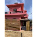 Two-story pink house with a decorative balcony, wooden slatted fence, and an open gate; entry stairs visible under a small porch against a blue sky.